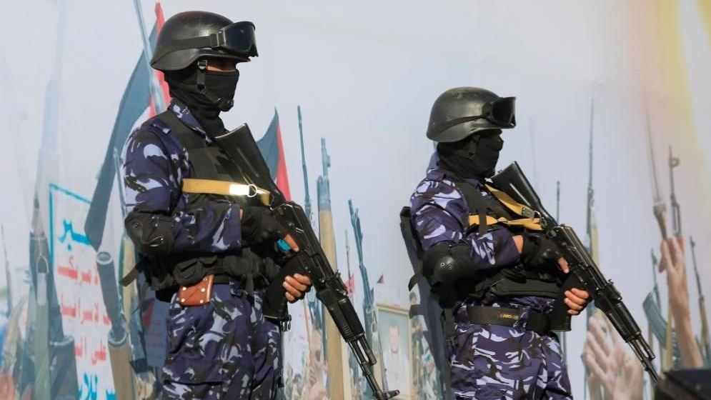 Two soldiers with guns in blue camouflage standing guard in front of a big mural that has many pictures of guns on it.
