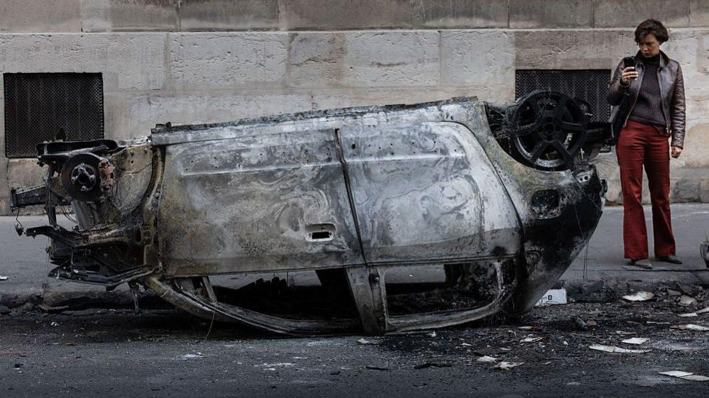 A woman takes a photo of the wreckage of a burnt‑out car in central Paris